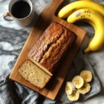 A freshly baked golden-brown banana bread loaf sliced on a wooden cutting board, accompanied by ripe bananas, sliced bananas, and a cup of coffee