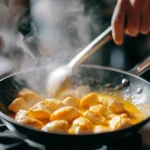 Chicken pieces simmering in a vibrant yellow ají amarillo sauce in a skillet, with steam rising as a hand stirs using a spoon.