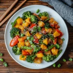 A vibrant and colorful stir-fry featuring kabocha squash, broccoli, red bell peppers, and snap peas, garnished with sesame seeds and green onions, served on a white plate with wooden chopsticks beside it.