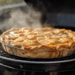 A golden-brown homemade apple pie with a flaky crust, steaming fresh inside a pellet grill, infused with a subtle smoky flavor.