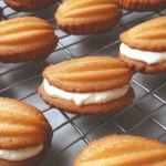 Golden-brown madeleine cookies filled with creamy whipped filling, cooling on a wire rack.