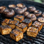 Marinated tofu cubes and portobello mushrooms grilling over an open flame, with a rich, smoky char and crispy seared edges.